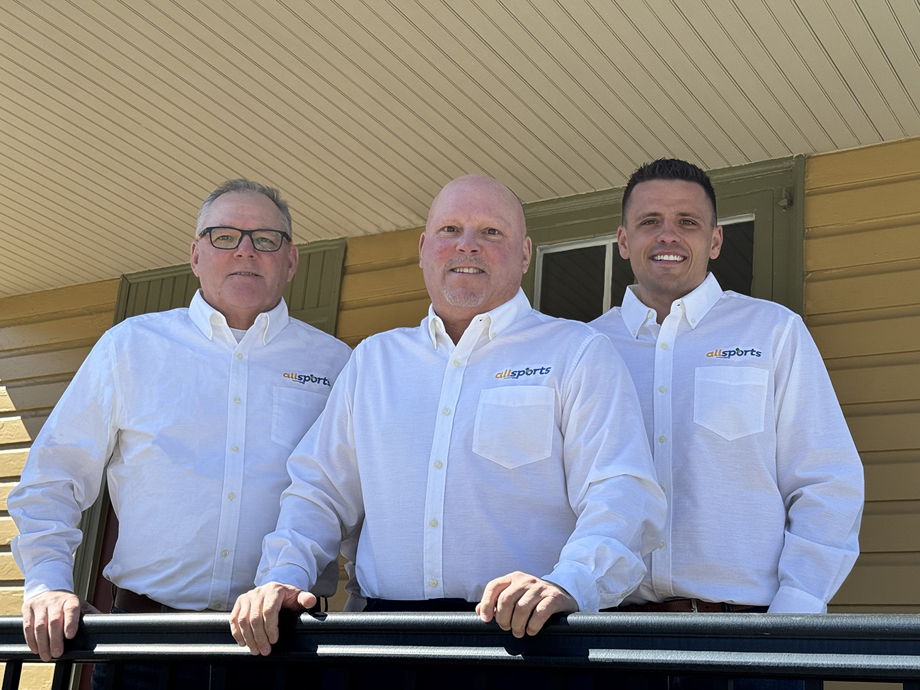 Three AgPro team members in branded white button-downs standing together outside, representing the leadership behind a trusted sports construction company partnership.